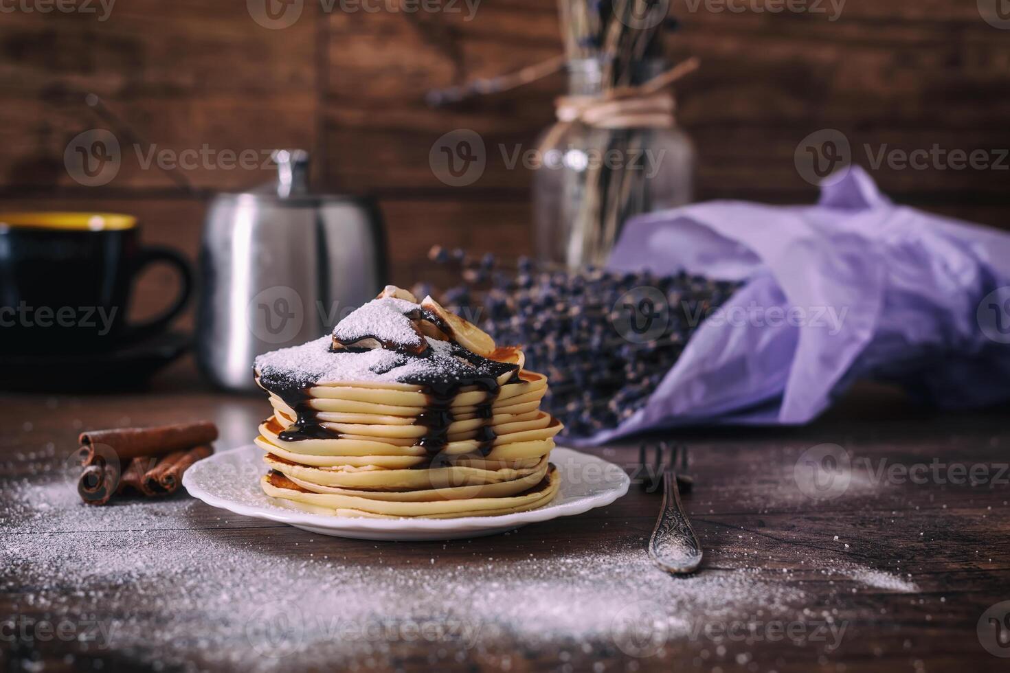 Stack of delicious pancakes with chocolate, honey, slices of banana and castor sugar on plate on wooden background, cinnamon sticks and bunch of lavender. photo