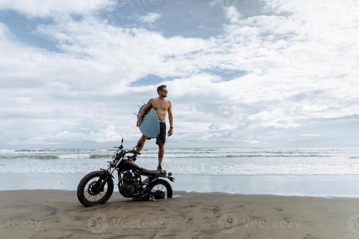 male person looking for inspiration while standing on sportive motorbike. holding surfboard in hands. Young man surfer enjoying recreation near to ocean. photo