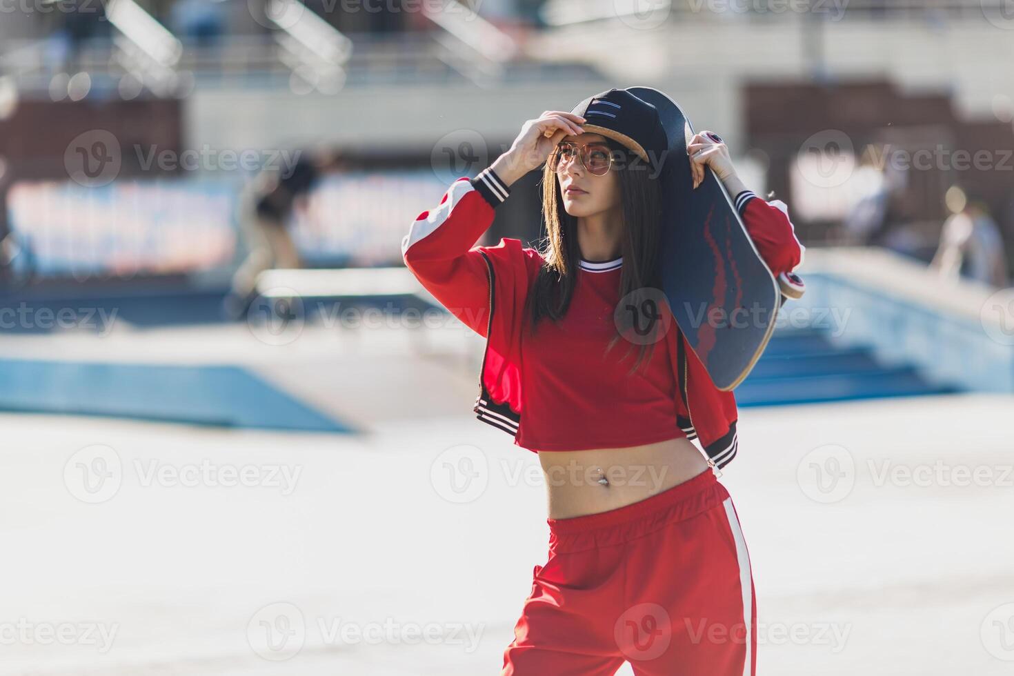 Portrait of young female wearing black hat, red clothing skateboarder holding her skateboard. Woman with skating board looking at camera outdoors. photo