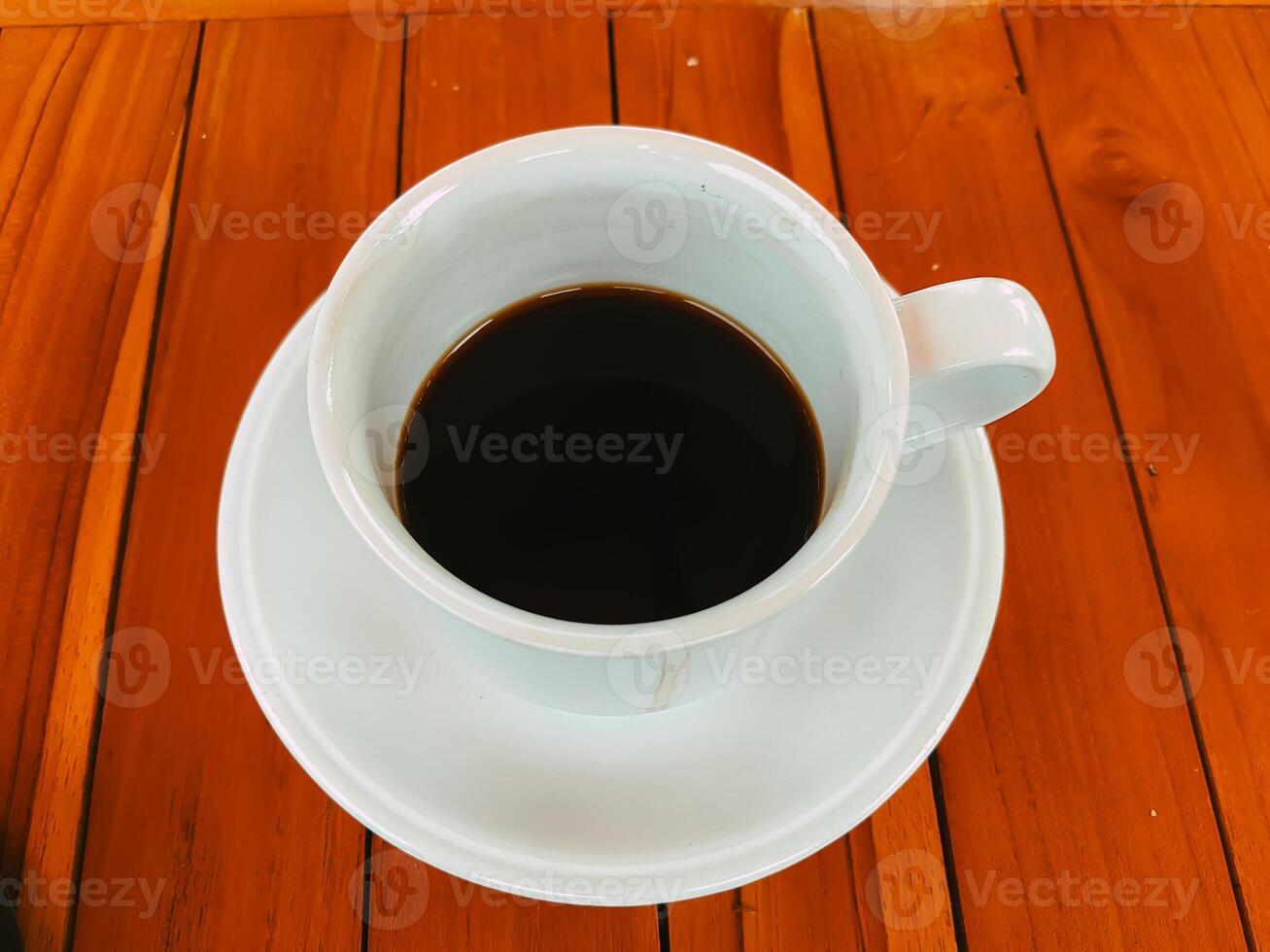 A white ceramic cup filled with black coffee sits on a rich, wooden table. The warm tones of the wood contrast with the sleek, smooth surface of the cup photo
