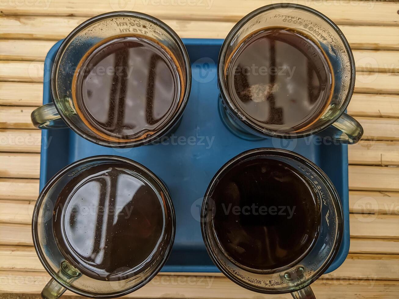 A top view of four glass mugs filled with dark coffee placed symmetrically on a blue tray atop a rustic bamboo surface. The natural lighting highlights reflections on the coffee surface. photo