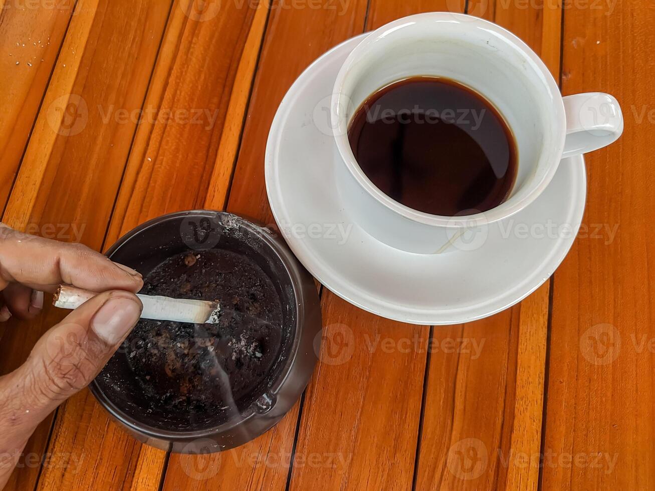 A hand holding a cigarette over an ashtray beside a cup of black coffee on a wooden table captures a moment of calm photo