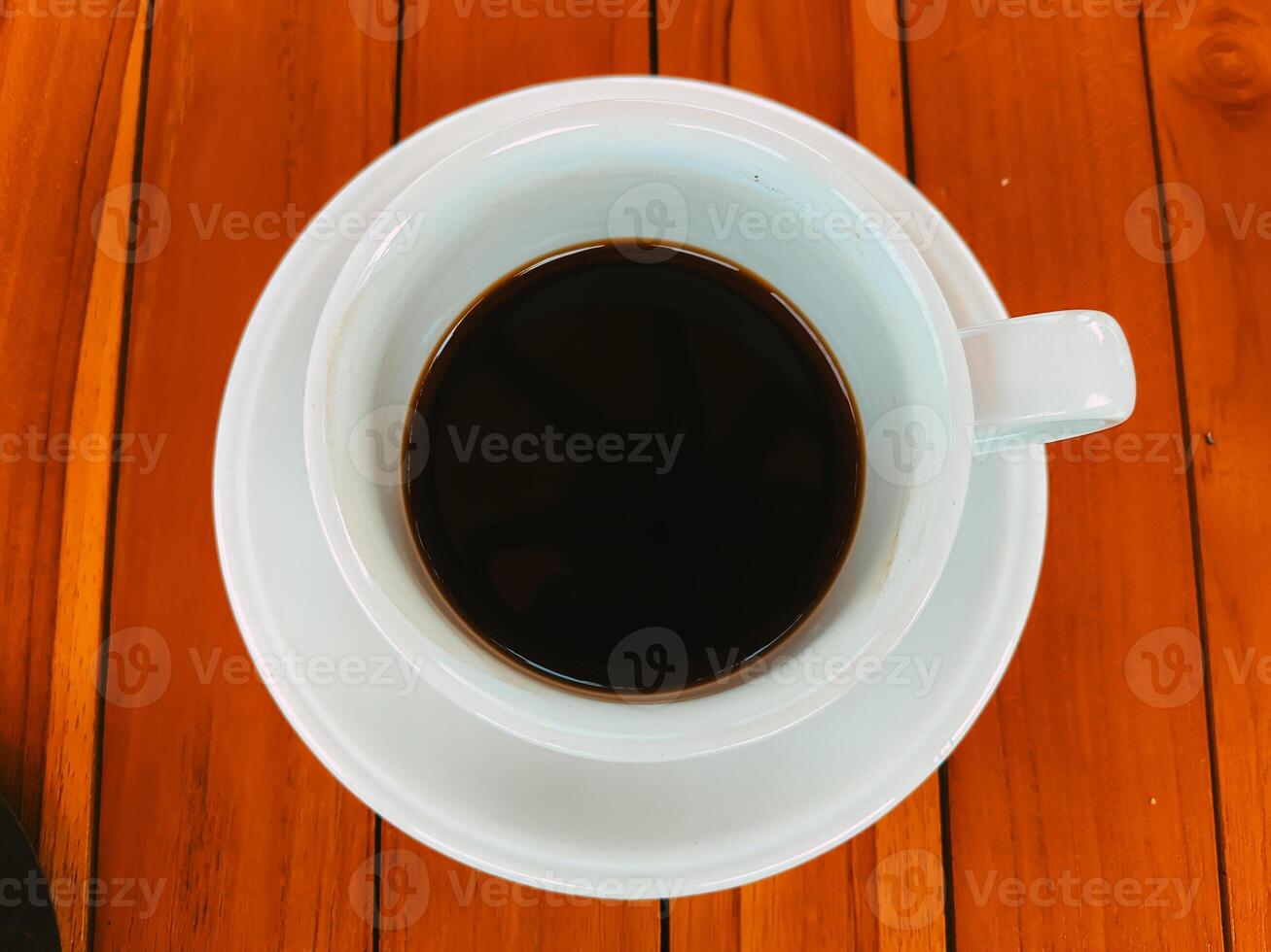 A top-down view of a white coffee cup filled with black coffee on a wooden table. The coffee is dark and strong, and the cup is the only object in the frame. photo