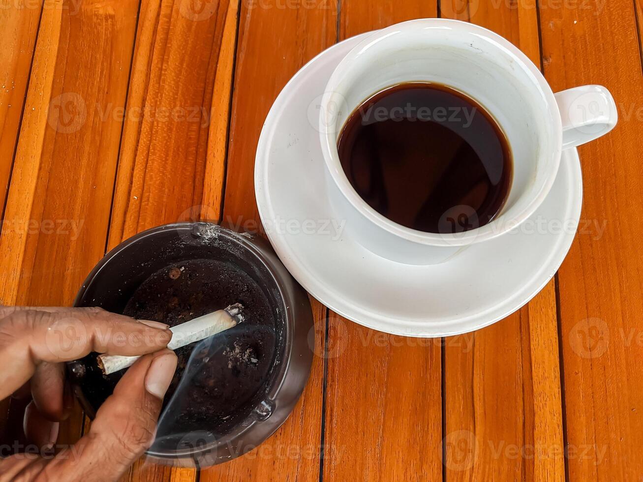 A close-up image of a hand holding a cigarette over an ashtray beside a cup of black coffee on a wooden table, capturing a moment of relaxation and contemplation. photo