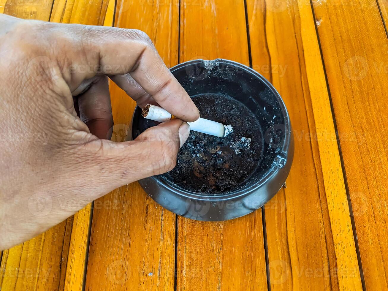 A person hand extinguishes a cigarette in a black ashtray. The wooden table contrasts with the ashtray, creating a vivid scene that represents the end of a smoking moment photo