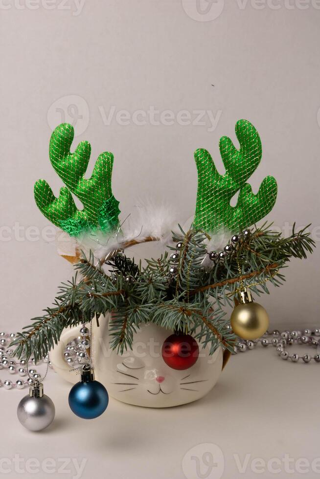 An alarm clock, a hoop with deer antlers and branches of a Christmas tree decorated with toys are placed in a cute white cup in the shape of a sleeping cat's head photo