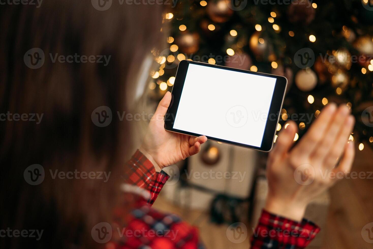 Woman holding tablet with blank screen in front of Christmas tree, festive bokeh lights in background, ready for holiday call or virtual greeting. photo