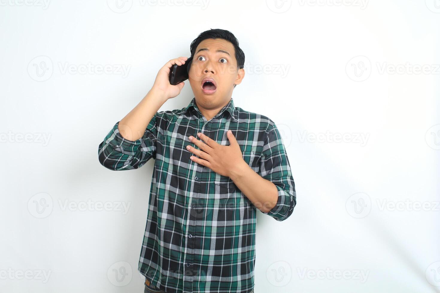 shocked asian man touching chest while talking on smartphone wearing casual shirt isolated on white background photo