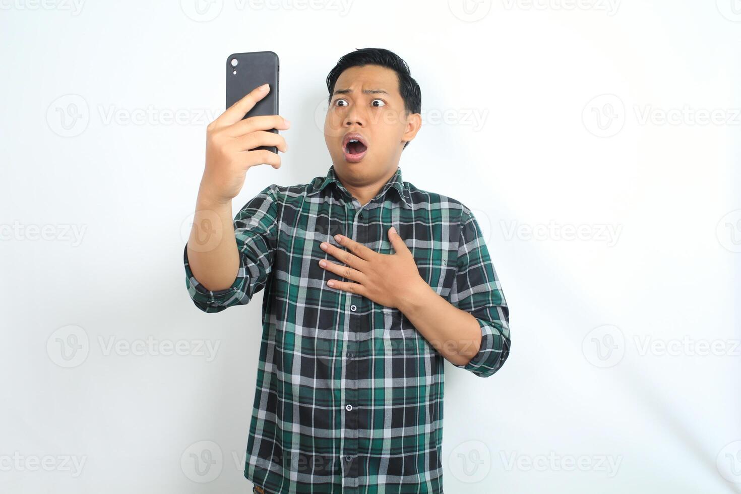 asian man shocked while looking at smartphone, touching chest, isolated on white background photo
