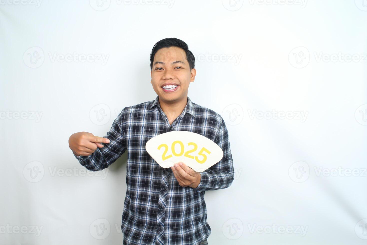 carefree asian man pointing to 2025 new year eve greeting card with smiling face isolated on white background, male celebrating new year eve photo