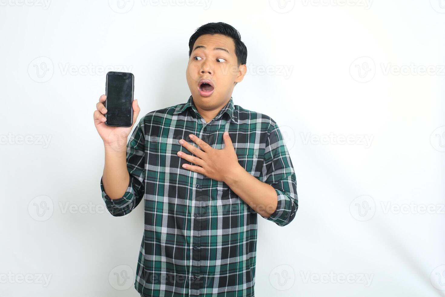 isolated asian man showing phone screen with shocked expression, touching chest, wearing casual shirt photo