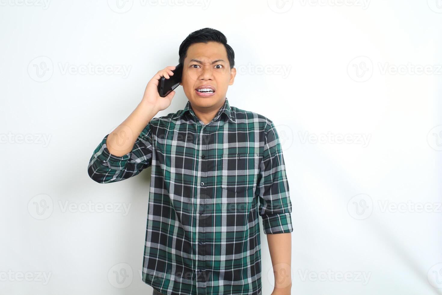 unhappy asian man wearing casual plaid shirt looks angry while talking on cellphone isolated on white background photo