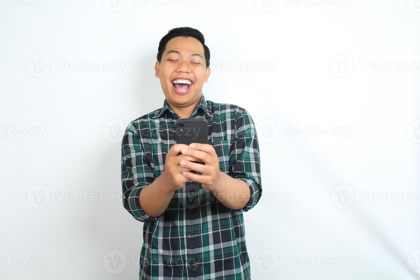 carefree asian man wearing casual plaid shirt laughing while watching on smartphone isolated on white background photo