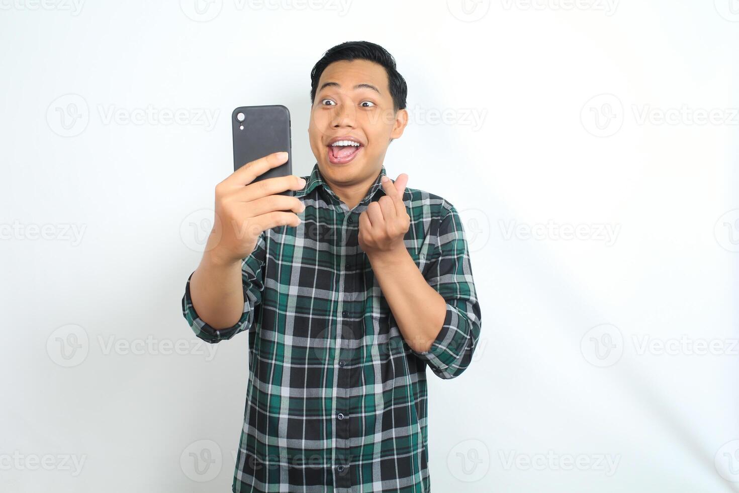 surprised asian man holding cellphone and giving heart love shape isolated on white background, wearing casual green shirt photo