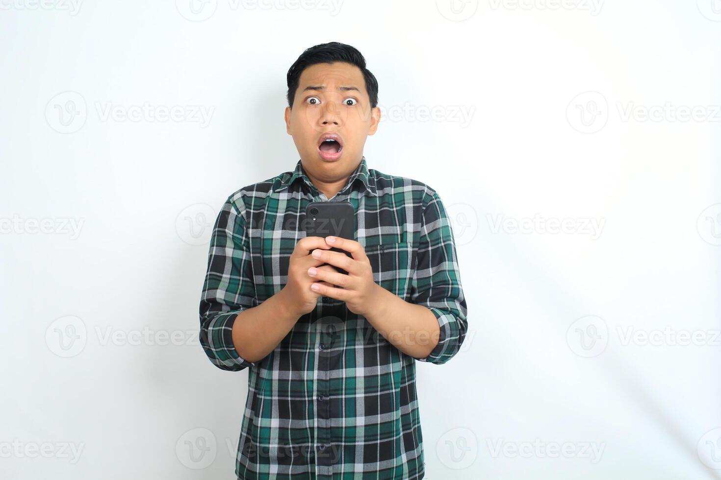 asian man holding smartphone show shocked face expression, looking at camera, wearing casual shirt, isolated on white background photo