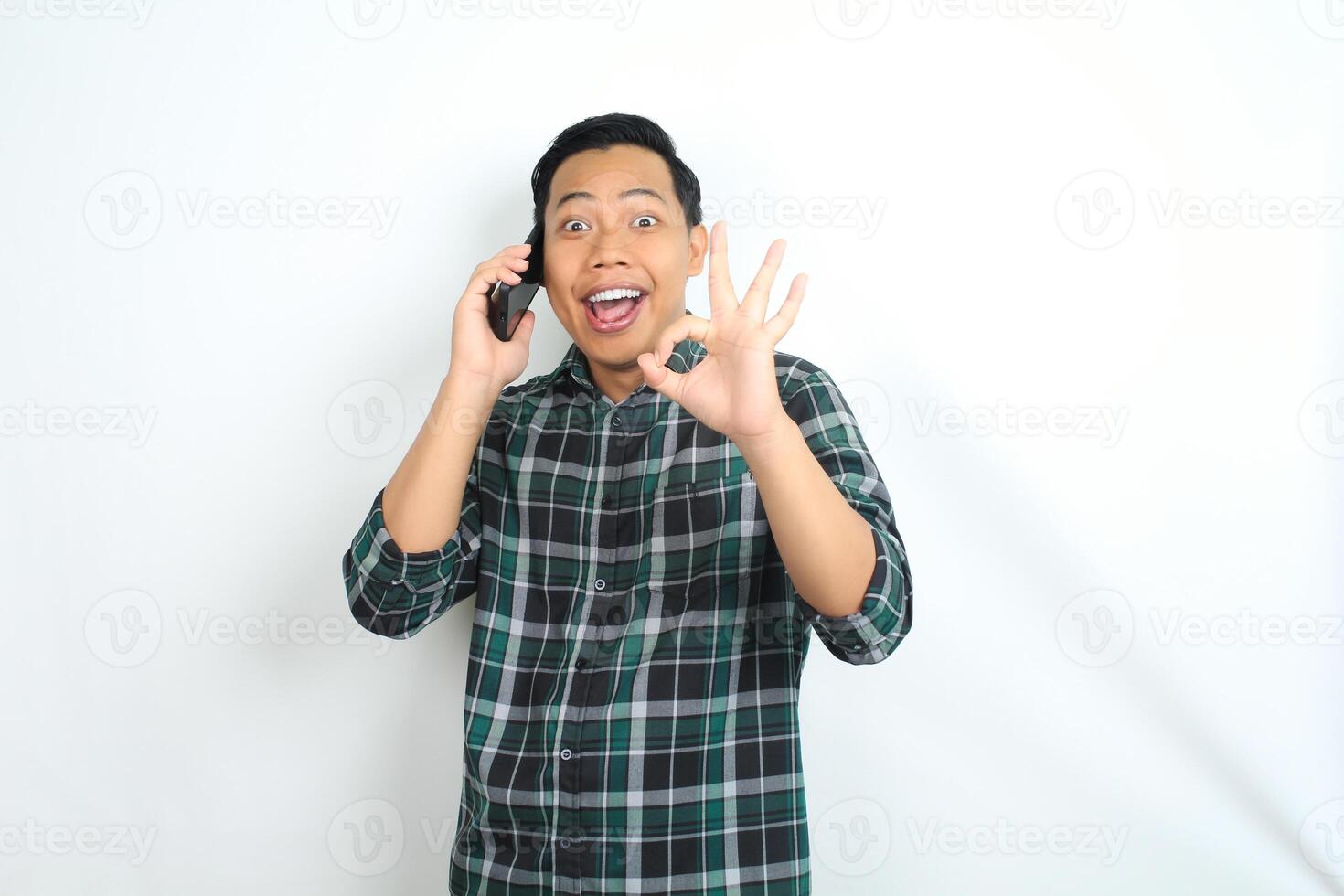 yes gesture, asian man give okay sign with surprised face while talking on smartphone isolated on white background, wearing casual green shirt photo