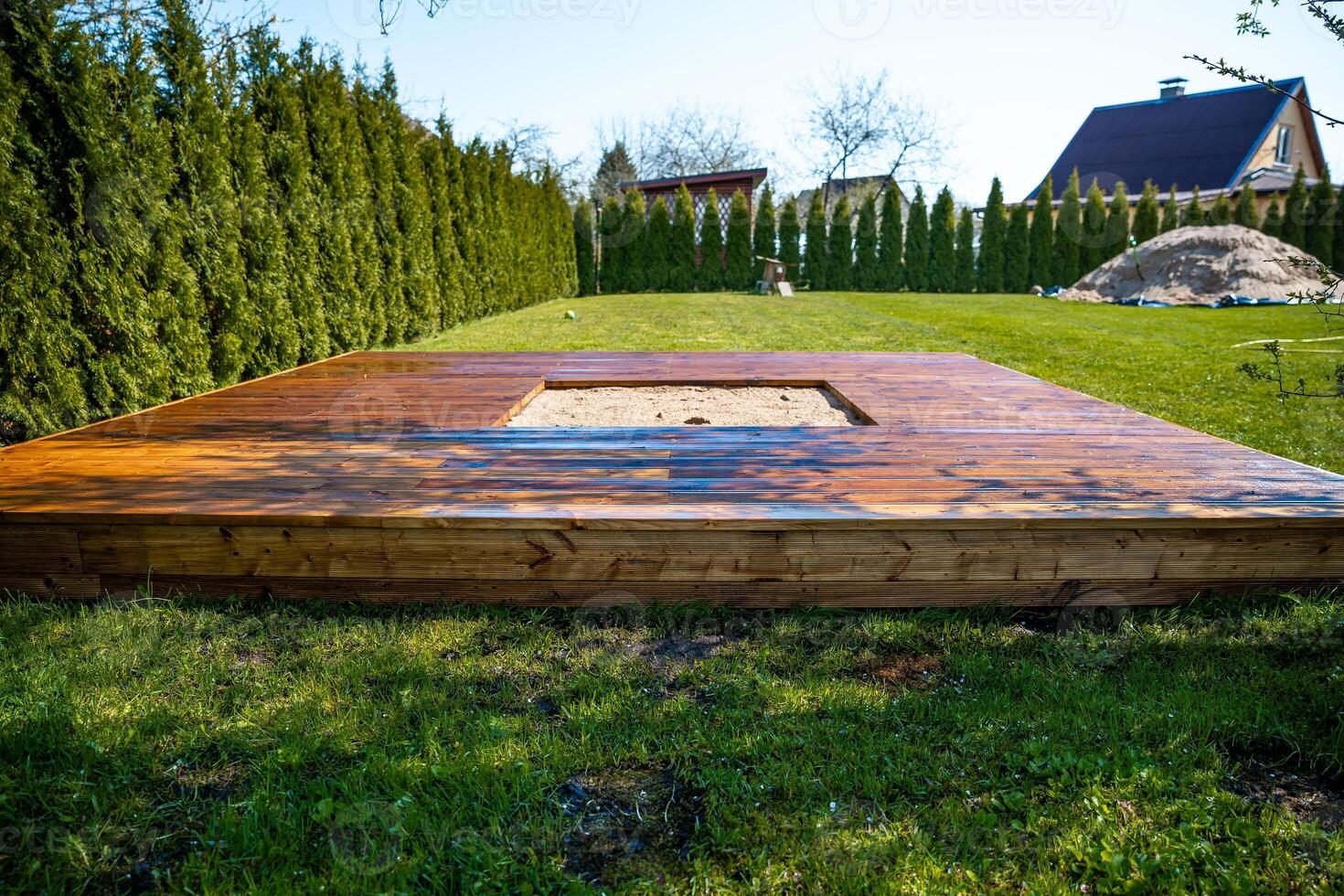 Freshly stained wooden deck with a central sandpit in a backyard surrounded by trees and bushes photo