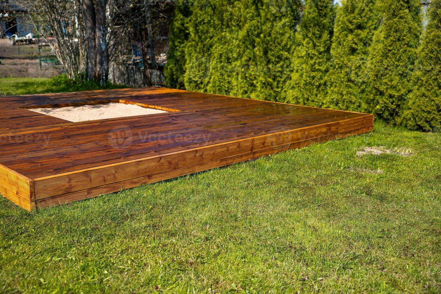 Freshly stained wooden deck with a central sandpit in a backyard surrounded by trees and bushes photo