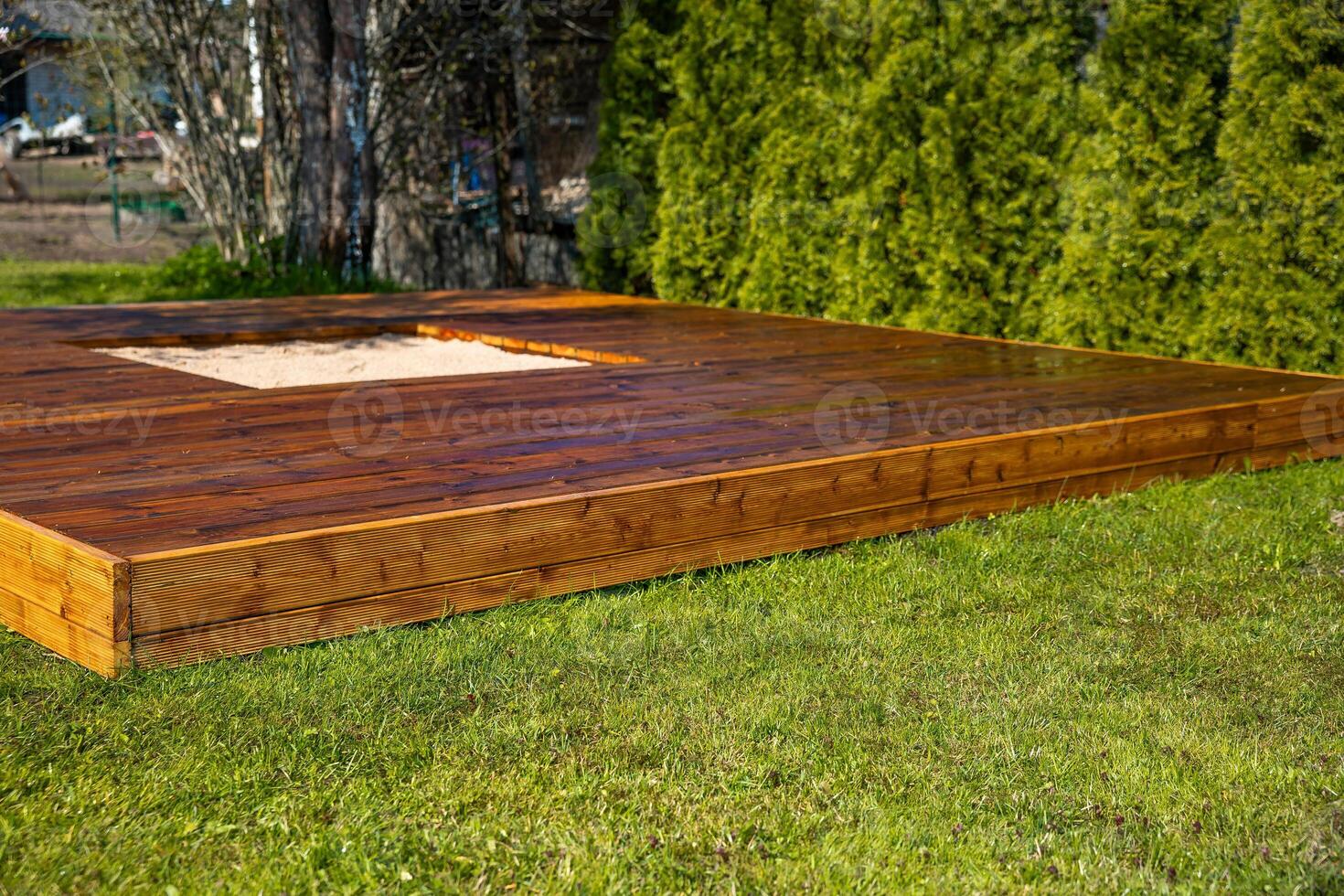 Freshly stained wooden deck with a central sandpit in a backyard surrounded by trees and bushes photo