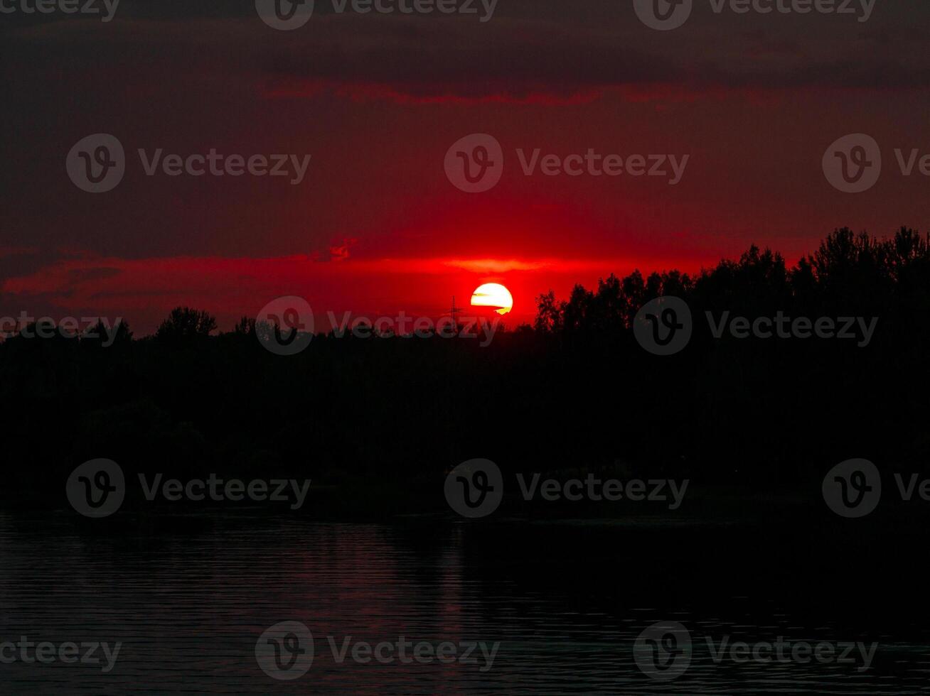 Vivid sunset over a dark forest, with the bright orange sun just above the treetops, casting a warm glow over the landscape. photo