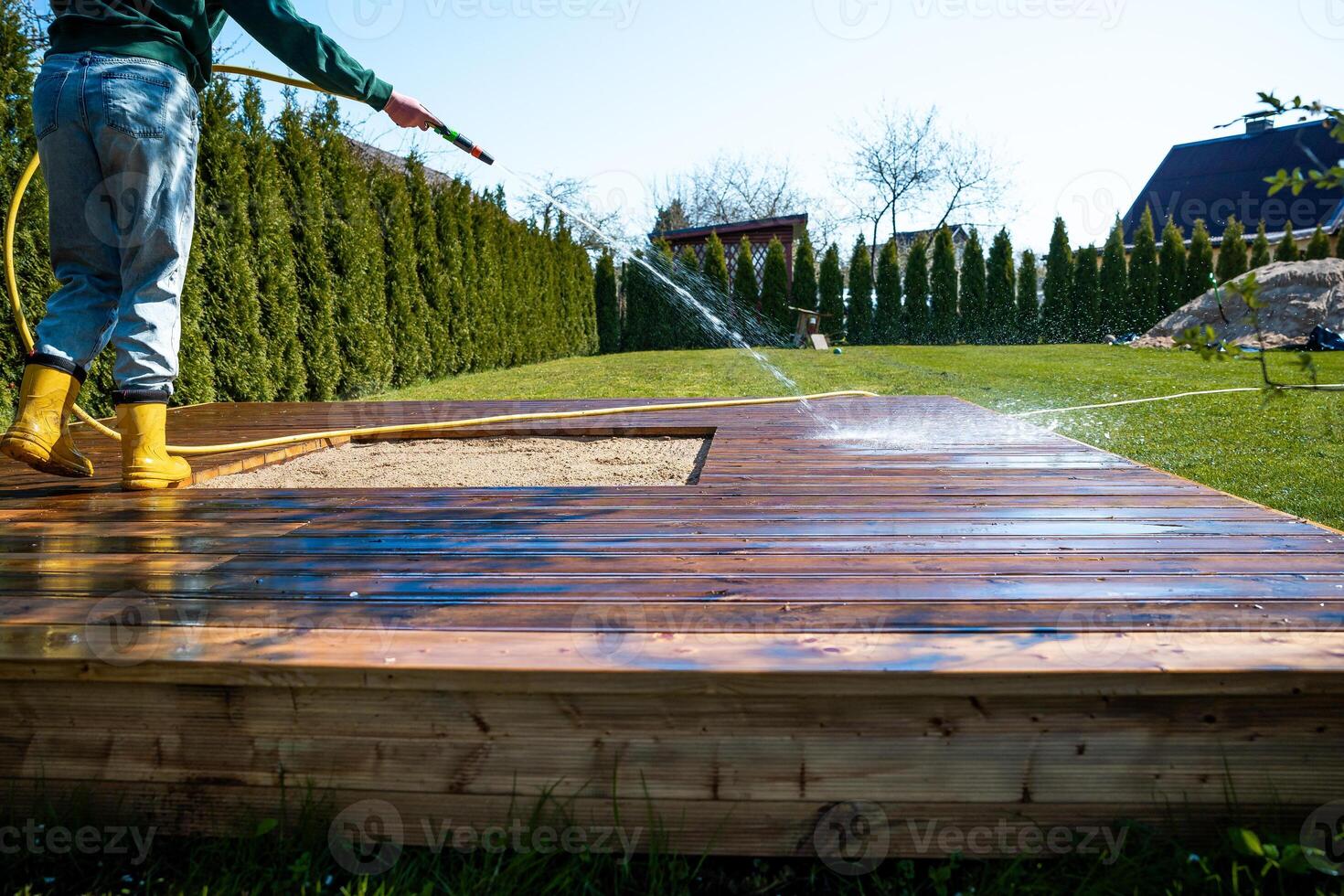 Young woman cleaning a wooden deck terrace with a hose in a sunny backyard photo