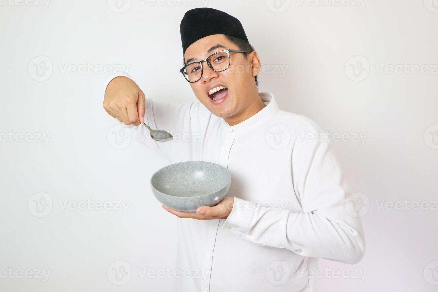 Asian muslim man wants to empty bowl and holding spoon with funny expression, isolated white background photo