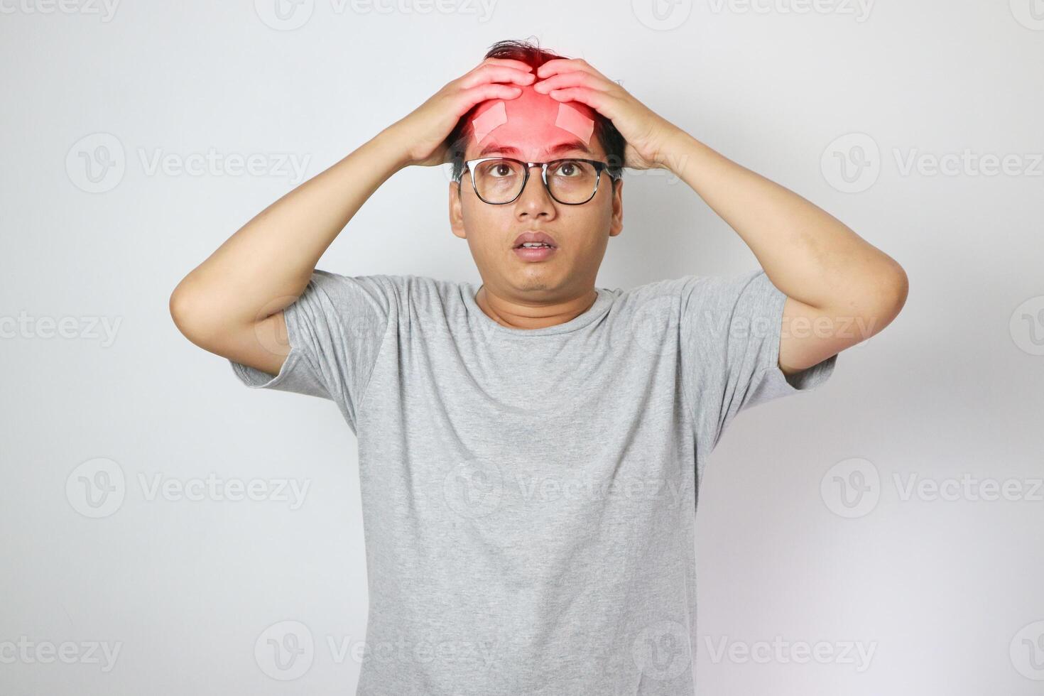 A man in a grey shirt holds his head with a pained expression, indicating a headache or discomfort. The plain light blue background emphasizes his distress and highlights his posture and expression photo