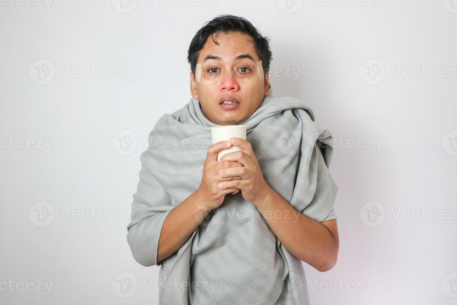 Young Asian man covers himself with grey blanket and shows cold expression while holding glass and drinking hot tea or coffee. Isolated on white background photo