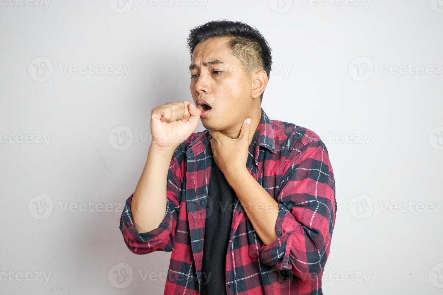 Young Asian man in casual t-shirt coughing into his fist while holding his chest, indicating discomfort or illness. Studio shot on plain background capturing his casual outfit photo