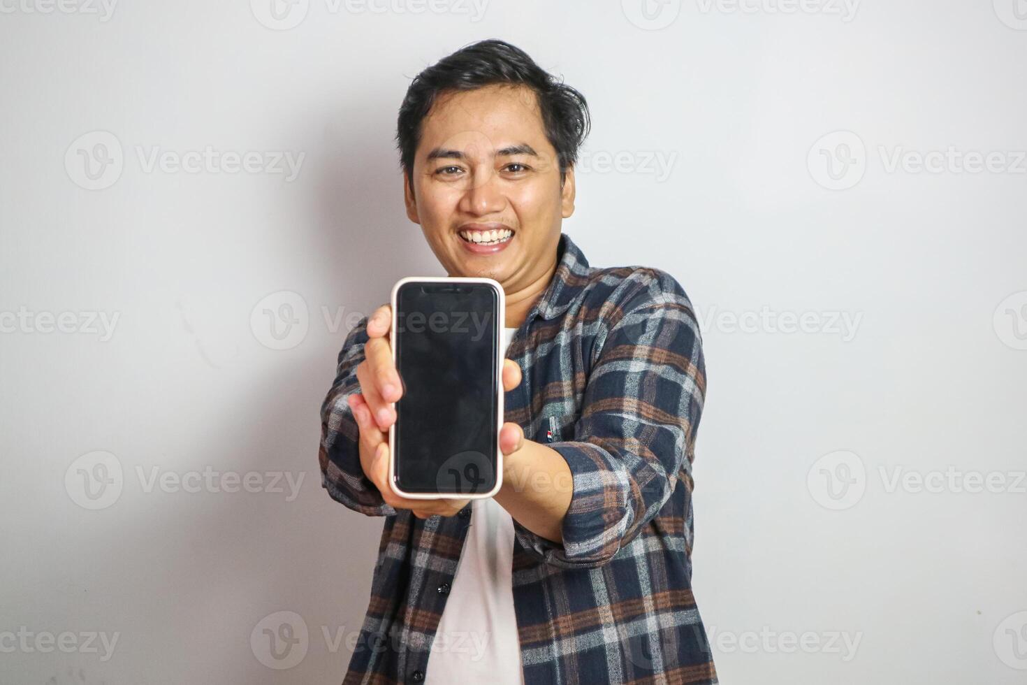 Happy young Asian man in casual shirt holding smartphone with blank screen isolated on white background photo