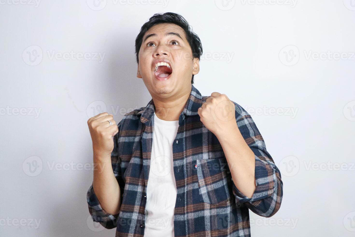 A young Asian man in a casual shirt pumps his fists in excitement and smiles broadly against a solid yellow background. This image captures the essence of celebration and triumph photo