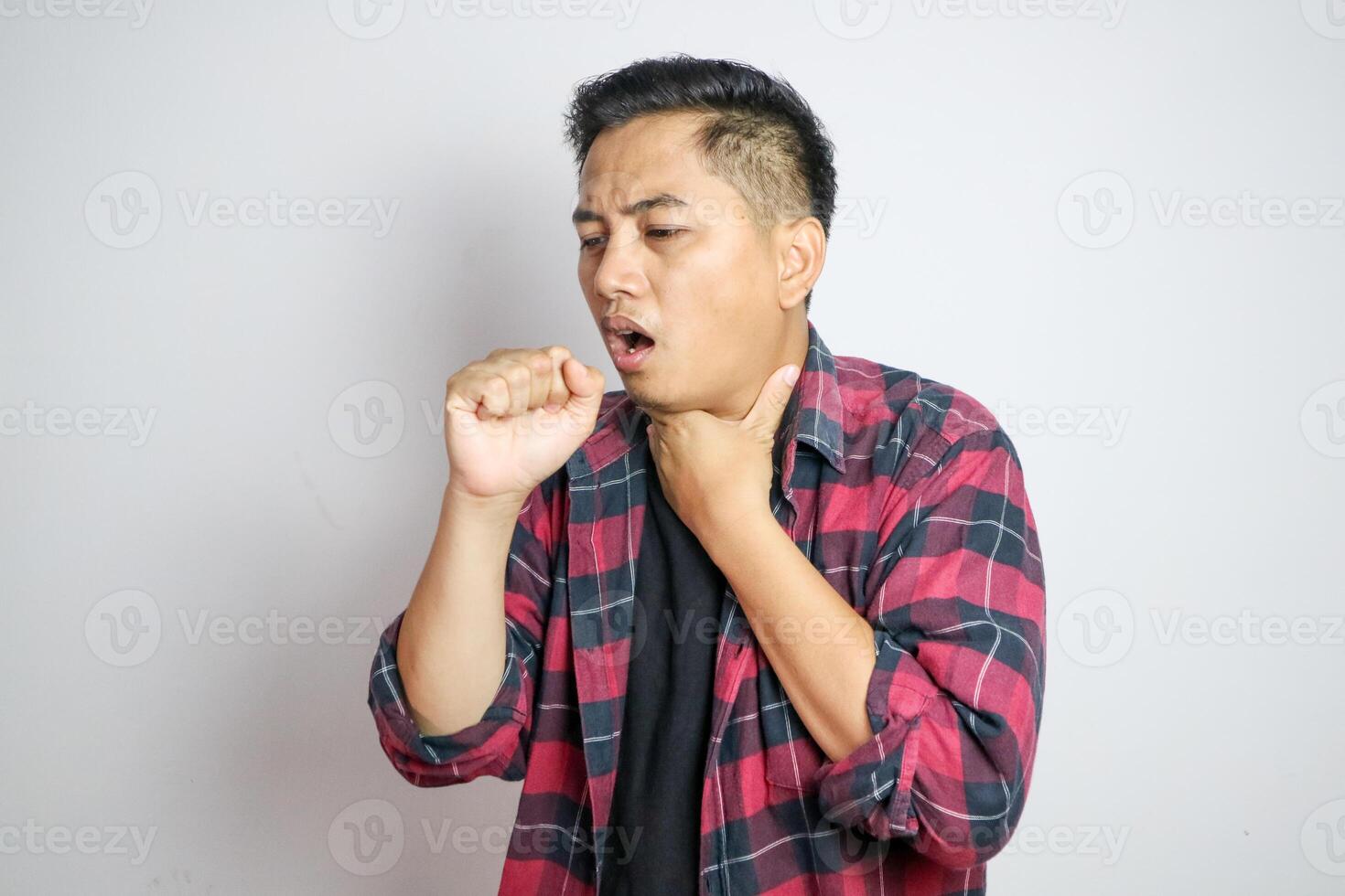 Young Asian man in casual t-shirt coughing into his fist while holding his chest, indicating discomfort or illness. Studio shot on plain background capturing his casual outfit photo