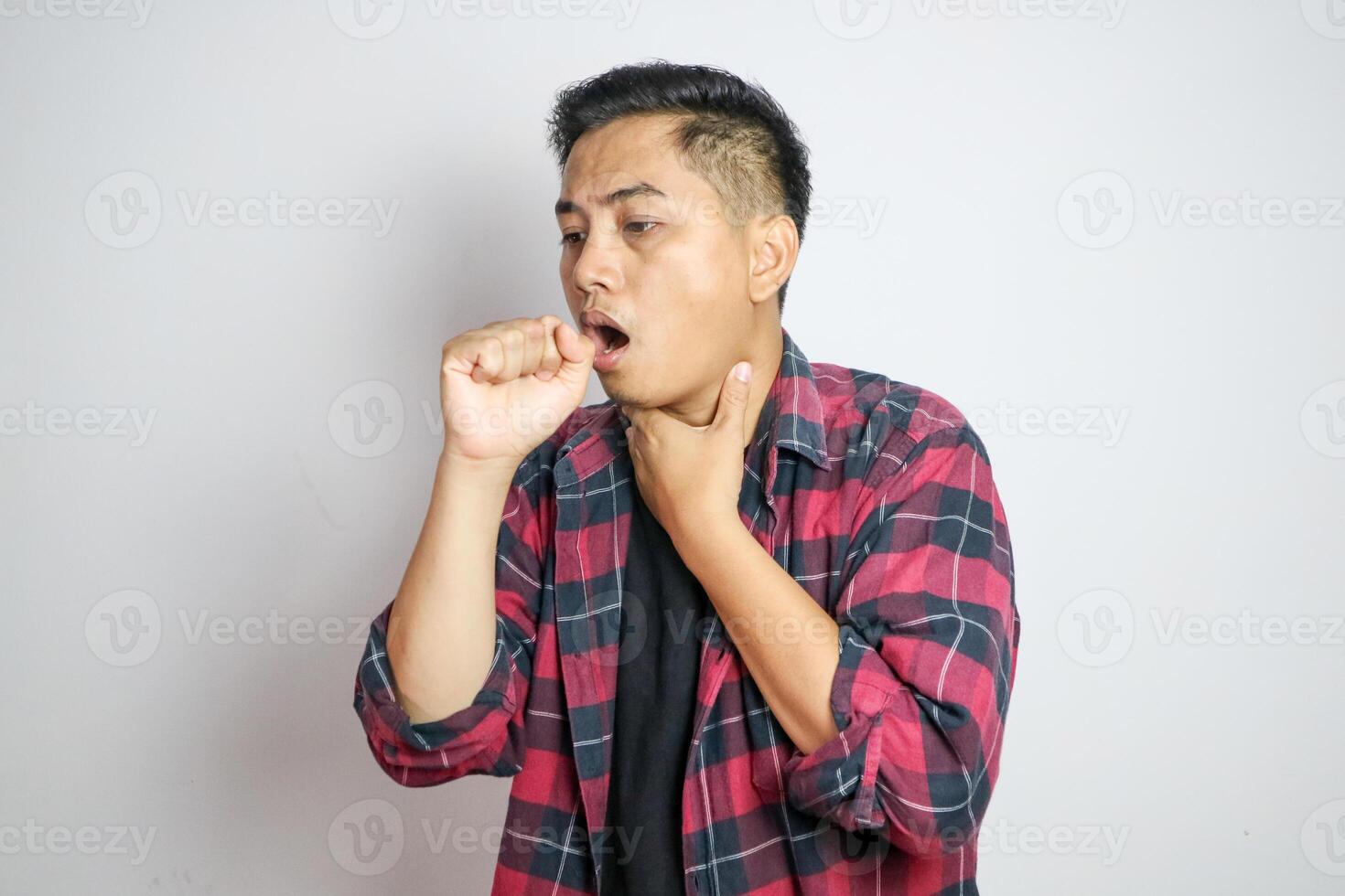 Young Asian man in casual t-shirt coughing into his fist while holding his chest, indicating discomfort or illness. Studio shot on plain background capturing his casual outfit photo