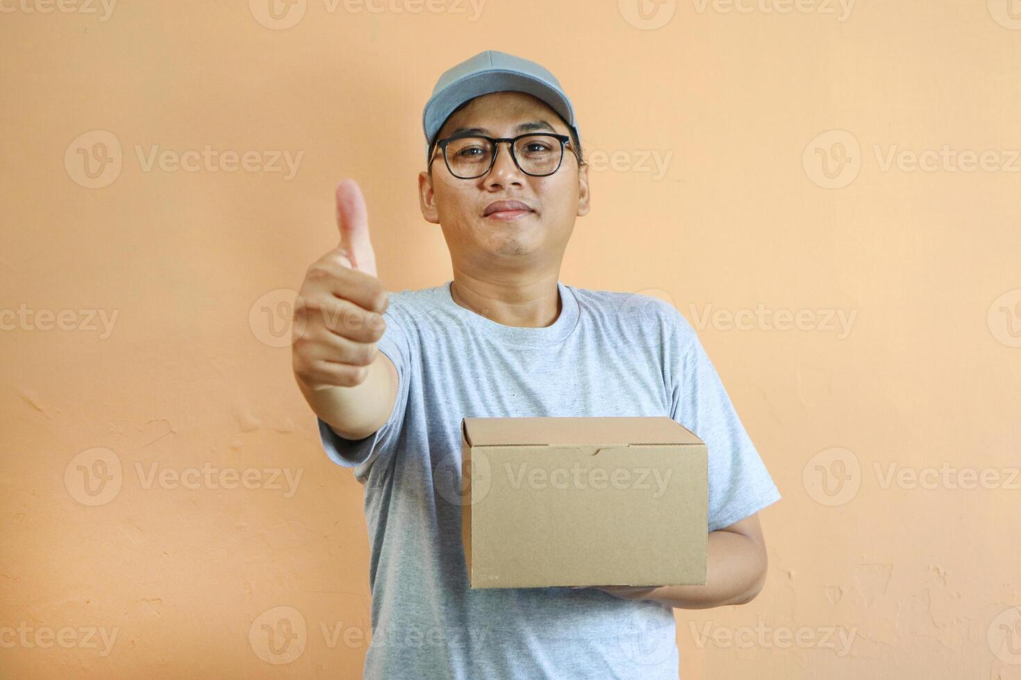 Satisfied young Asian man in grey cap t-shirt uniform, employee work as dealer courier carrying package box, showing thumb up sign isolated on yellow background. Professional Delivery service concept photo