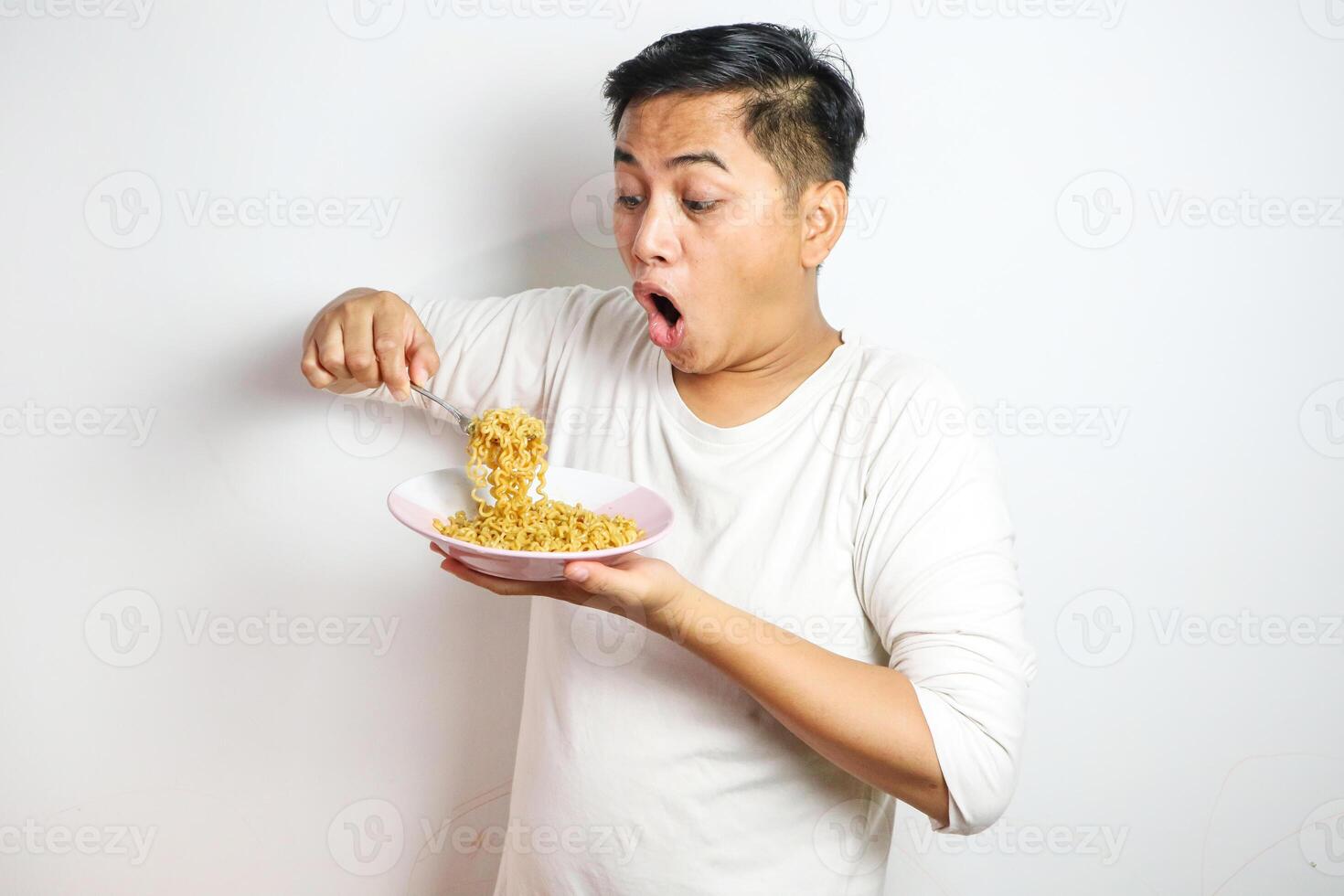 Asian Man grasping and lifting up Noodles Hungrily from Bowl using fork. Hungry Asian Man Devours Delicious Noodles photo