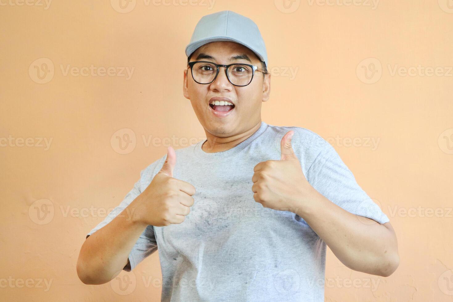 Satisfied young Asian man in grey cap t-shirt uniform, employee work as dealer courier carrying package box, showing thumb up sign isolated on yellow background. Professional Delivery service concept photo