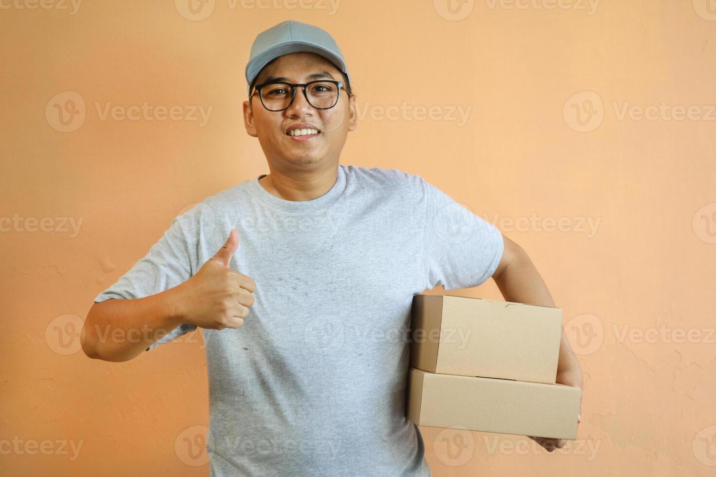 Satisfied young Asian man in grey cap t-shirt uniform, employee work as dealer courier carrying package box, showing thumb up sign isolated on yellow background. Professional Delivery service concept photo