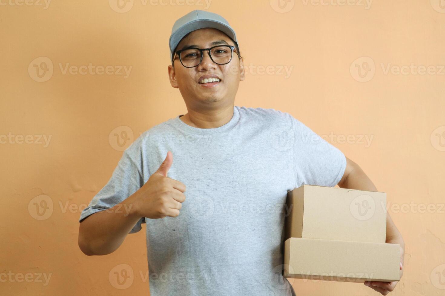 Satisfied young Asian man in grey cap t-shirt uniform, employee work as dealer courier carrying package box, showing thumb up sign isolated on yellow background. Professional Delivery service concept photo