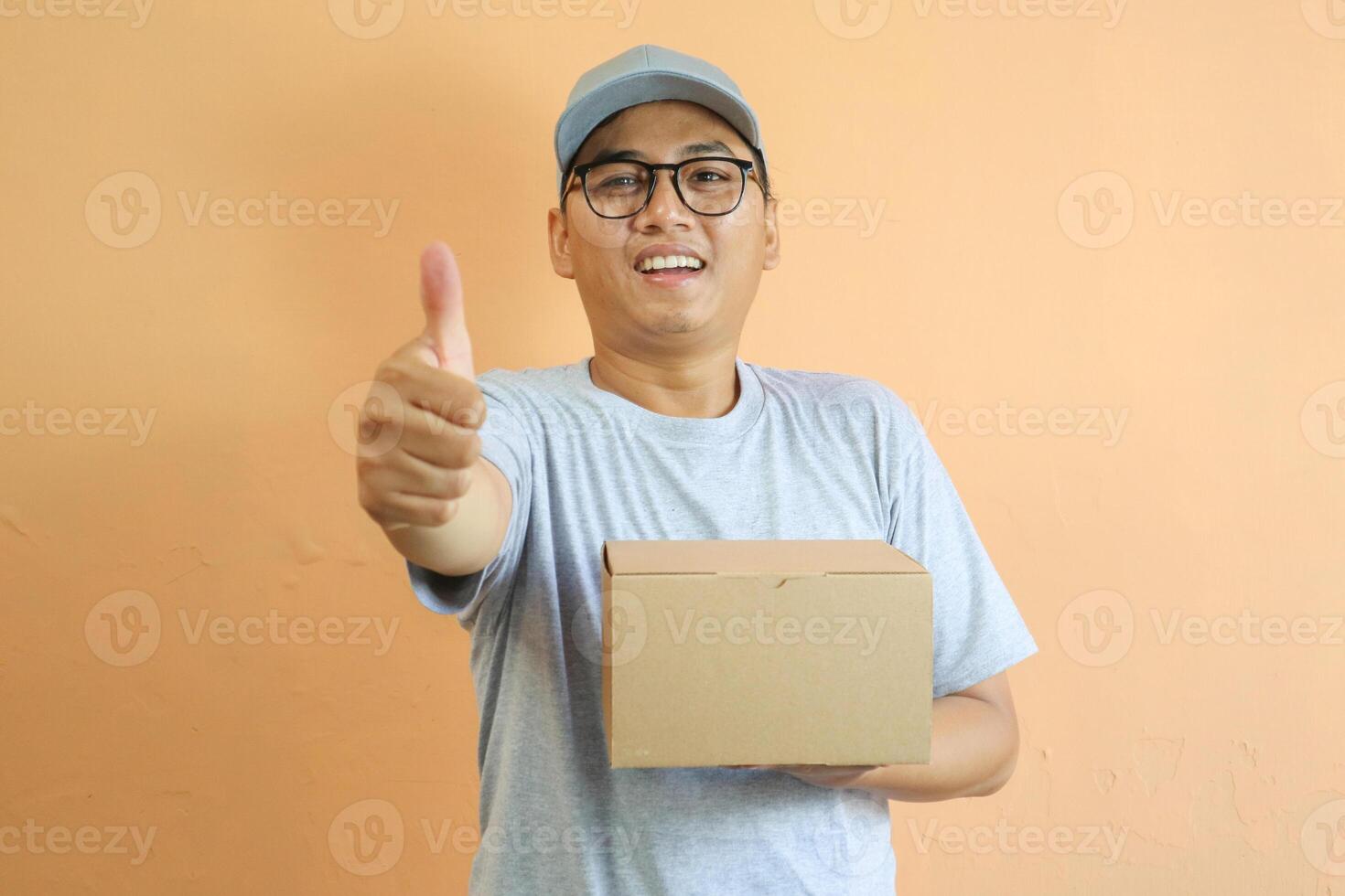 Satisfied young Asian man in grey cap t-shirt uniform, employee work as dealer courier carrying package box, showing thumb up sign isolated on yellow background. Professional Delivery service concept photo