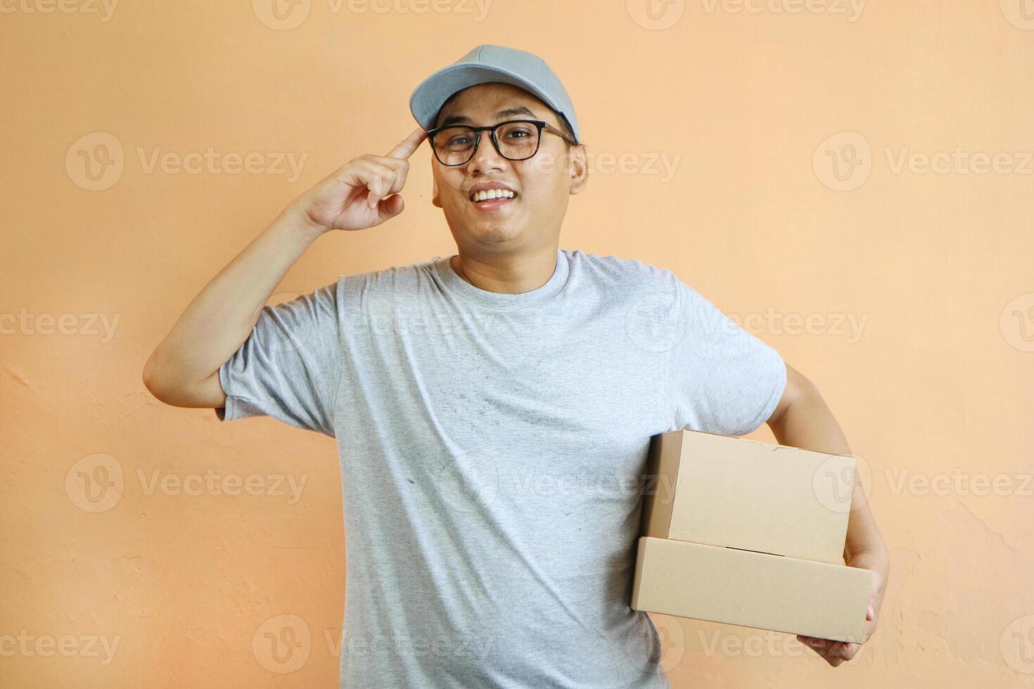 Pensive Asian delivery man or courier looking to the side at empty copy space, with his hand on his chin in thinking gesture, while carrying cardboard box package parcel, isolated on white background photo