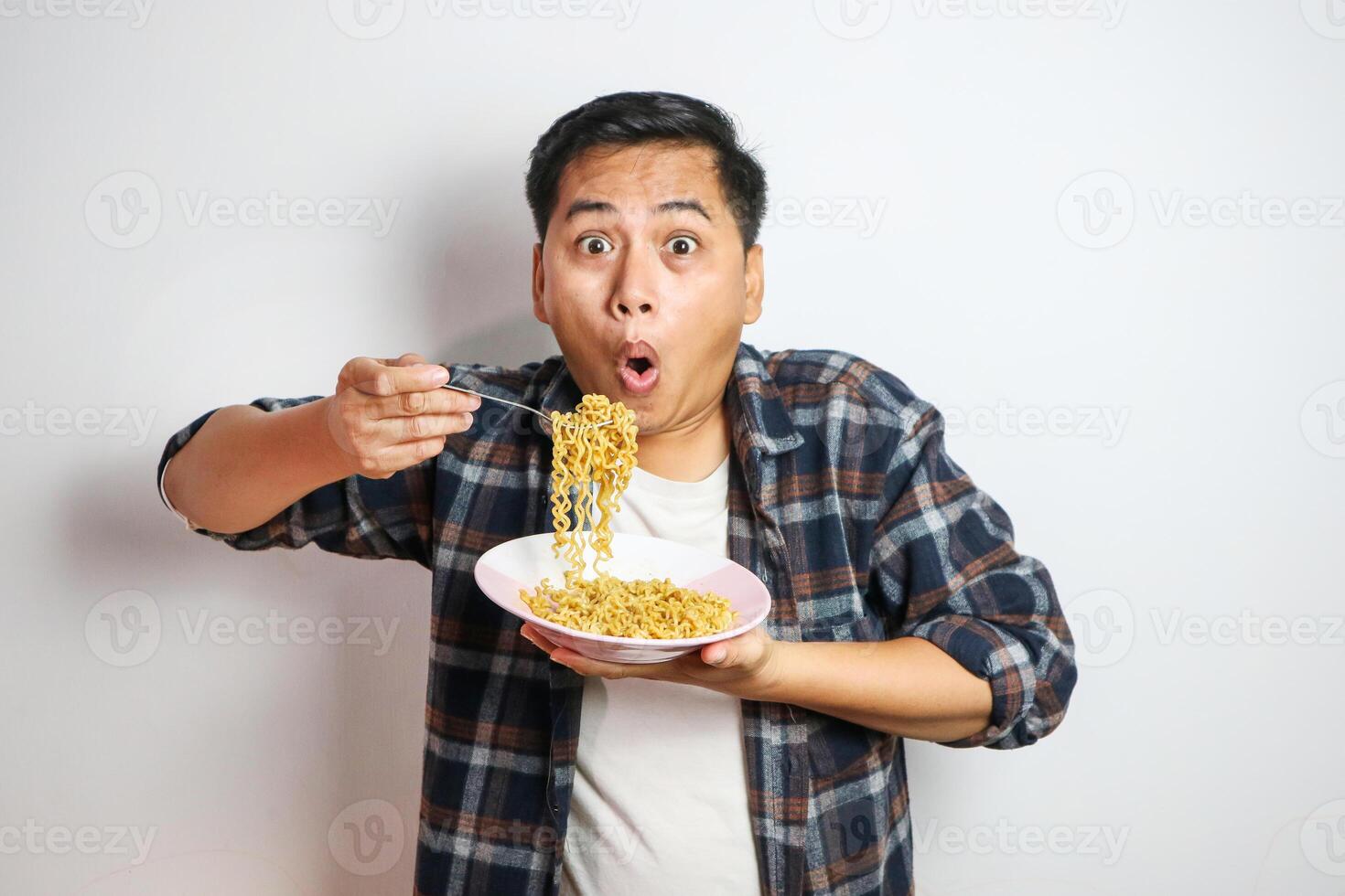 An Asian man in a casual shirt eagerly eats noodles from a plate, his expression filled with delight. The plain white background focuses on his enjoyable dining experience. photo
