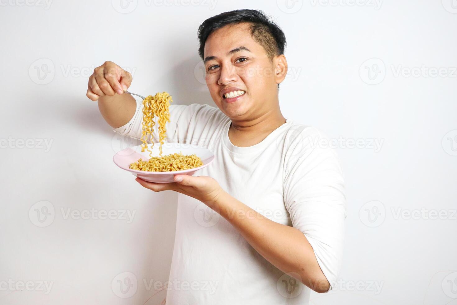A happy, euphoric Asian young man in a tshirt eating instant fried noodles using fork isolated over a white background. stylish asian man with peek a boo hair eats instant noodle photo