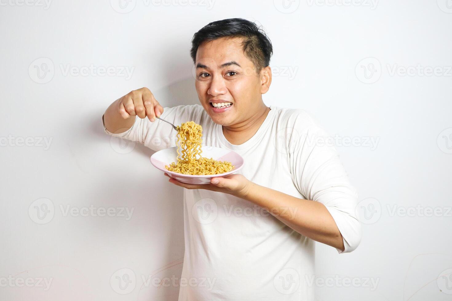Young Asian man in a white t-shirt enthusiastically eating instant noodles from a bowl with a fork. Studio shot on a plain background, highlighting his joyful expression and casual fashion photo