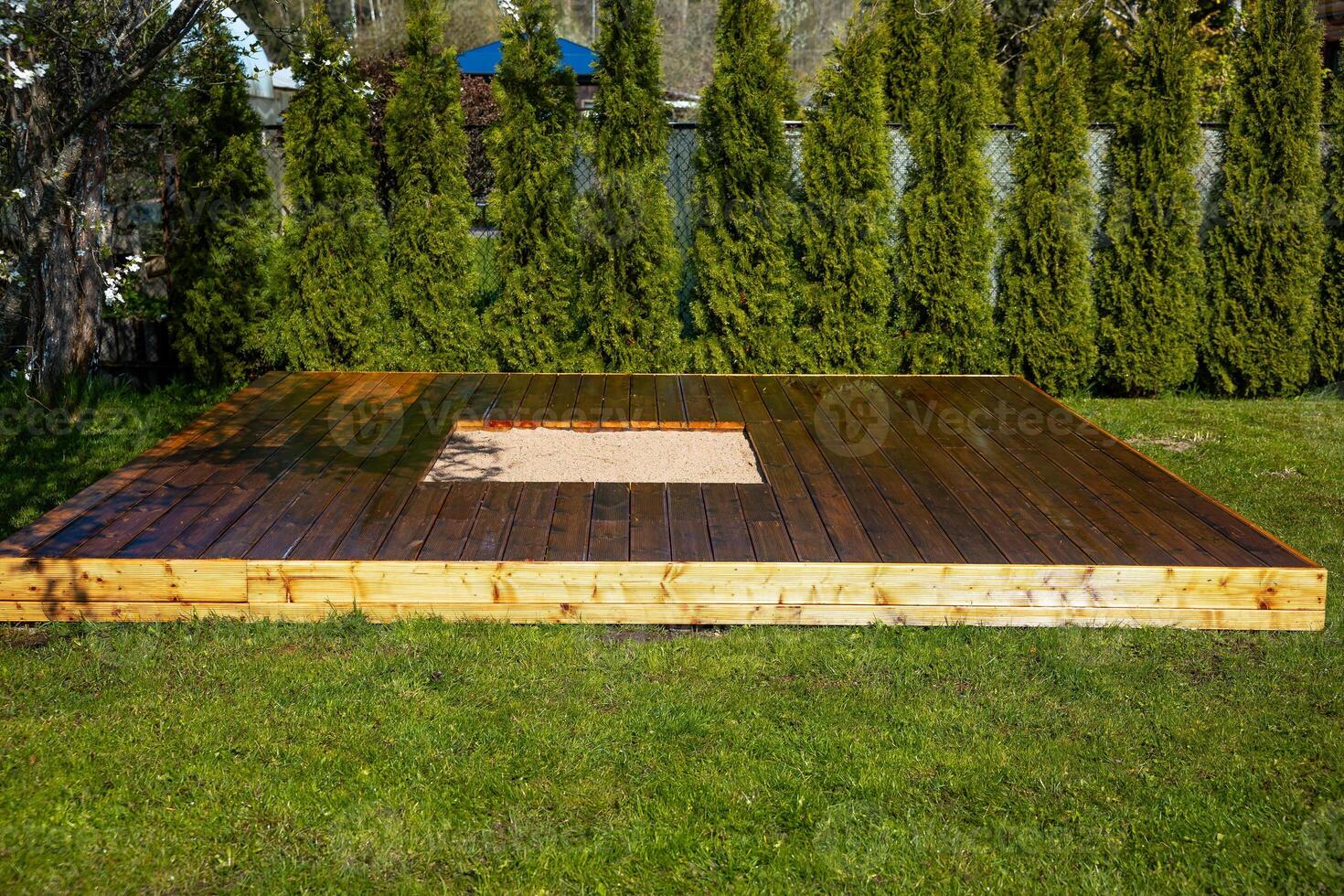 Freshly stained wooden deck with a central sandpit in a backyard surrounded by trees and bushes photo