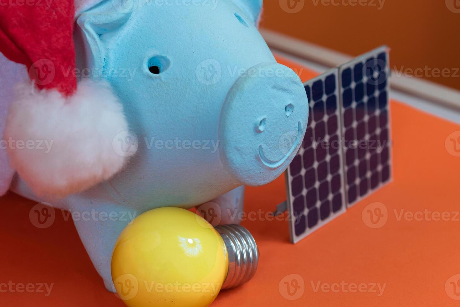 Piggy bank coins with Santa Claus christmas hat, photovoltaic solar panel and lamp isolated on orange background. Concept image. photo