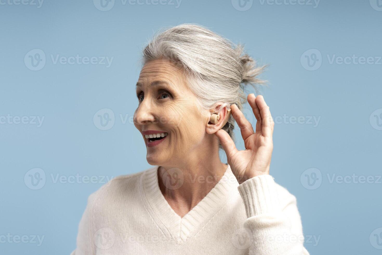 Portrait of deaf senior gray haired woman having hearing aid in ear, isolated on blue background photo