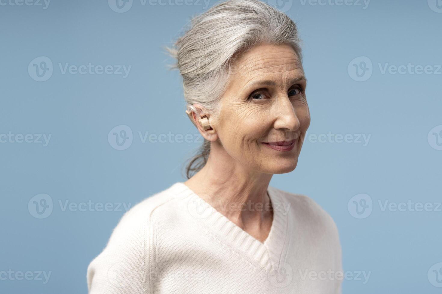 Portrait of smiling, senior gray haired woman having hearing aid in ear, isolated on blue background photo