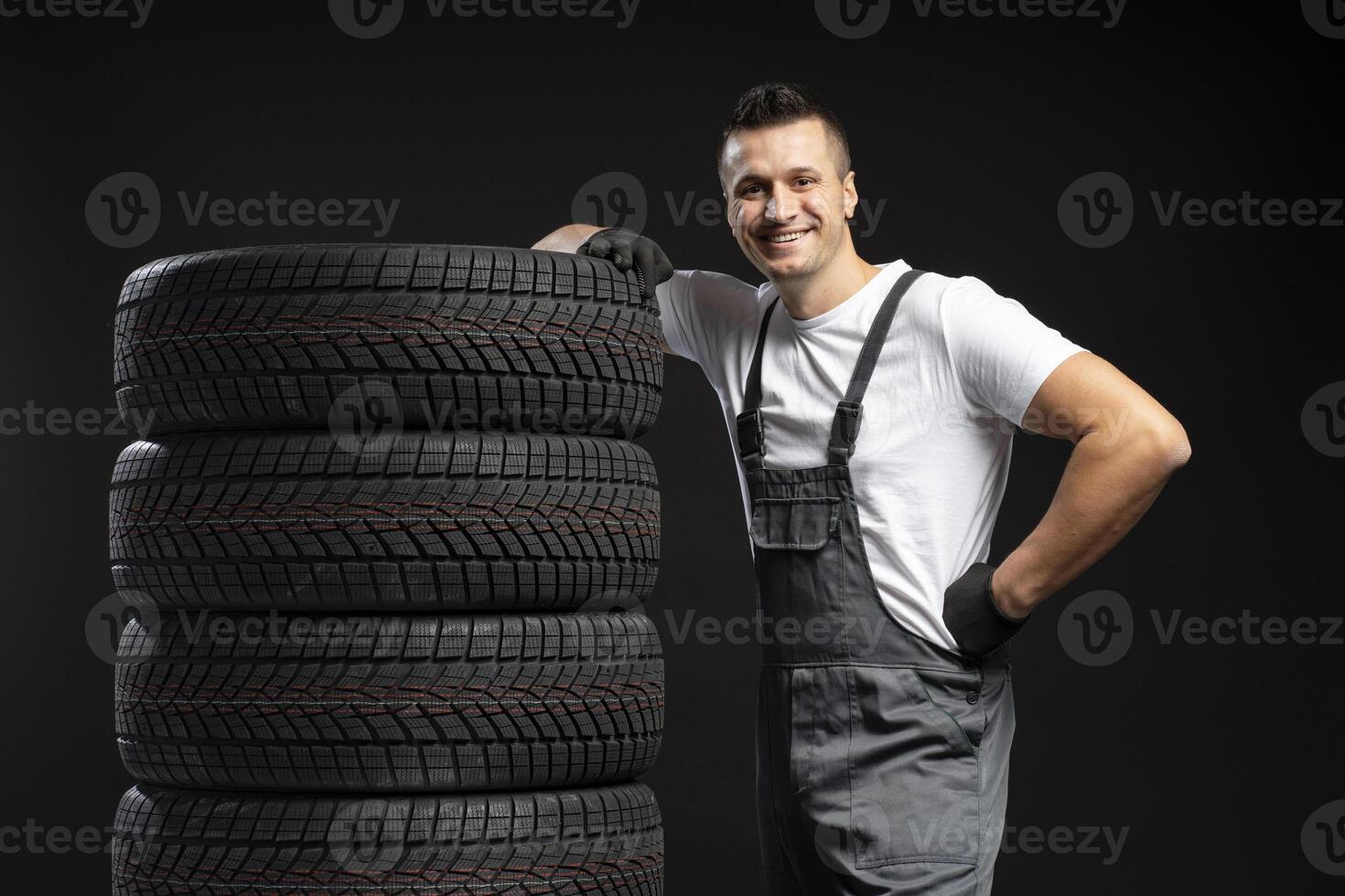 Mechanic leaning on stack of new winter tires posing on black background photo