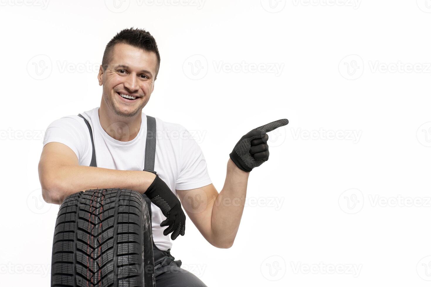 Smiling mechanic pointing aside while leaning on tire on white background photo