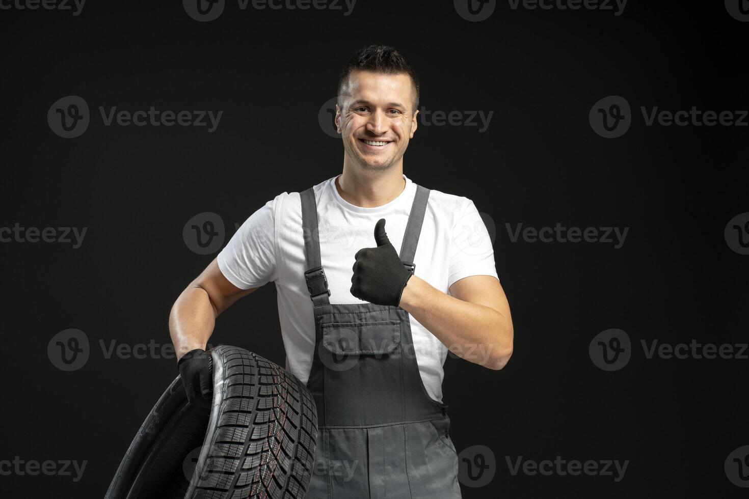 Mechanic holding car tire and showing thumbs up on black background photo